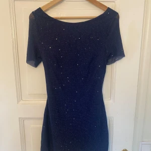 Mörkblå glittrig klänning - Säljer en superfin mörkblå klänning med glittriga detaljer. Den har en djup öppen rygg med knytband och korta ärmar. Perfekt för festliga tillfällen! ✨