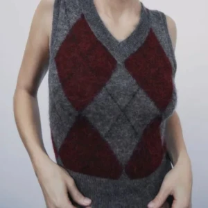 Grå och vinröd stickad väst - Säljer en snygg grå och vinröd stickad väst med ett klassiskt argyle-mönster. Västen har en v-ringad design och är perfekt för höst och vinter. Den är i bra skick och passar perfekt för en stilren och varm look.