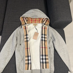 Burberry Check Zip Hoodie - Helt oanvänd, fick den som present men passar tyvärr inte. Har fått lega hemma.  Grå zip - Med huva   - Dragkedja fram, en känguruficka  - Långa ärmar och kontrasterande muddar  - Två fickor  - retail : 6000kr  Hör av er om ni har frågor! 