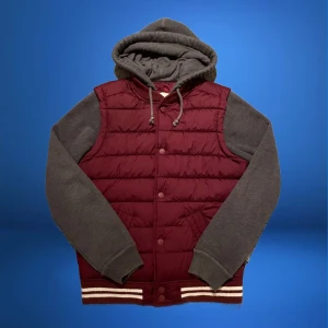 Vinröd pufferjacka med grå ärmar och huva - Säljer en snygg vinröd pufferjacka med grå ärmar och huva från hollister. Jackan har en quiltad design och stängs med knappar. Perfekt för höst och vinter, med en cool och avslappnad stil. Den har ribbstickade muddar och en skön passform. Perfekt för chill dagar eller en promenad i stan! Leverans sker med PostNord eller Instabox inom 48h!✅