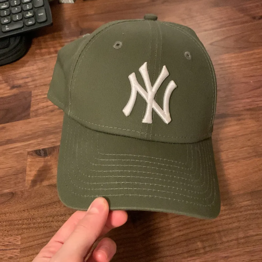 Säljer en snygg grön keps från New Era i modellen 9FORTY. Den har en broderad vit logga framtill och justerbar rem baktill för perfekt passform. Perfekt för både vardag och sportiga aktiviteter. Kepsen är i bra skick och redo för nya äventyr!. Övrigt.