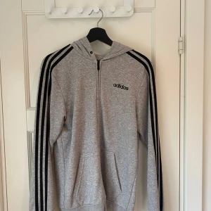 Grå hoodie från Adidas - Säljer en grå hoodie från Adidas med klassiska svarta ränder längs ärmarna och en dragkedja framtill. Den har en bekväm passform och är perfekt för både träning och vardag. Huvan ger extra mysfaktor och fickorna framtill är praktiska för småsaker. Perfekt för höst och vår!