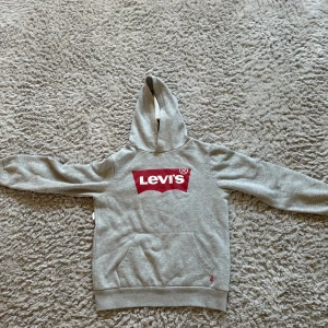 Grå hoodie från Levi's - Säljer en snygg grå hoodie från Levi's med deras klassiska röda logga på bröstet. Den är i bra skick och perfekt för både höst och vår. Hoodien har en känguruficka och ribbade muddar vid ärmslut och nederkant. Superbekväm och stilren!