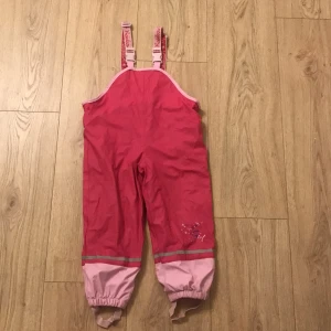 Rosa regnbyxor för barn - Säljer ett par supergulliga rosa hängselbyxor för barn. De är fodrade med mjukt material och har justerbara axelband. Perfekta för lek utomhus under höst och vinter. Byxorna har elastiska muddar vid bensluten för att hålla tätt mot kyla och väta. Och man kan minsa storleken vid midjan men knapparna som syns på bild 3 och 4!