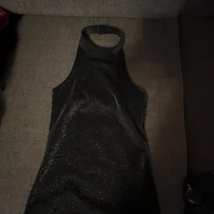 Svart glittrig klänning - Snygg svart glittrig klänning med hög krage och ärmlös design. Perfekt för festliga tillfällen när du vill glänsa lite extra. Klänningen har en tight passform som framhäver figuren. Materialet är mjukt och bekvämt, vilket gör den idealisk för en utekväll. Också stor storlek