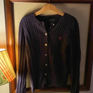 Mörkblå kofta från Ralph Lauren - Säljer en snygg mörkblå kofta från Ralph Lauren. Den är stickad med ett klassiskt kabelmönster och har knappar framtill. Perfekt för höst och vinter, med en liten röd logga på bröstet som ger en fin detalj. Passar både till vardags och lite finare tillfällen. Obs det är en barntröja. Står M 8-10 på tröjan.