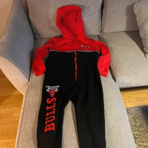 Chicago Bulls onesie i rött och svart - Säljer en supercool Chicago Bulls onesie i rött och svart. Den har en dragkedja framtill och en stor huva. Perfekt för att chilla hemma eller heja på ditt favoritlag! 🏀