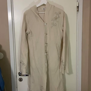 Beige kappa från Tippy - Säljer en elegant beige lång kappa från Tippy i storlek M. Den har fina broderade detaljer på ärmarna och axeln, samt knappar framtill. Perfekt för höst och vår! 🧥