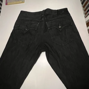 True Religion - Säljer ett par svarta jeans från True Religion i bra skick. De har en klassisk femficksdesign med snygga detaljer på bakfickorna och knappar. Perfekta för både vardag och fest! 🖤