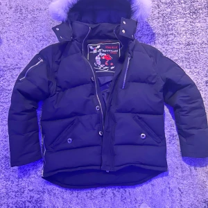 Moose Knuckles Jacka + Påse - Säljer en snygg svart pufferjacka från Moose Knuckles. Den har en fluffig pälsdetalj på huvan och dragkedja framtill. Perfekt för kalla vinterdagar! Jackan har flera fickor med knappar och en cool broderad logga på insidan. Den är i mycket bra skick och redo för nya äventyr.