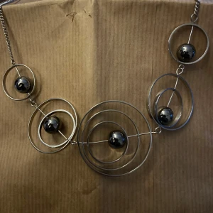 Unikt halsband med metallkulor - Säljer ett coolt halsband med en unik design av silverfärgade metallringar och svarta kulor. Perfekt för att ge en futuristisk touch till din outfit. Halsbandet har en kedja som gör det lätt att justera längden. Passar både till vardags och fest!