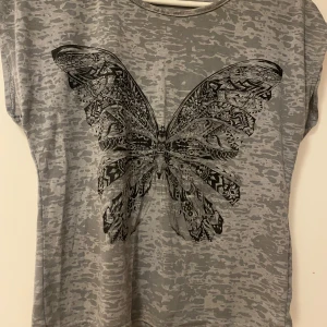 Grå t-shirt med fjärilsmönster - Säljer en snygg grå t-shirt med ett stort, detaljerat fjärilsmönster på framsidan. T-shirten har en avslappnad passform och korta ärmar, perfekt för vardagsbruk. Materialet känns mjukt och bekvämt mot huden. Passar bra till jeans eller shorts för en casual look.