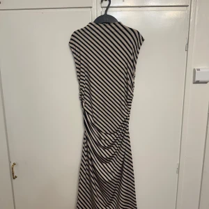 Randig klänning från H&M - Säljer en snygg randig klänning från H&M i storlek M. Klänningen har ett diagonalt randigt mönster i svart och beige, vilket ger en cool look. Den är ärmlös och har en draperad detalj i midjan som ger en smickrande passform. Perfekt för både vardag och fest!