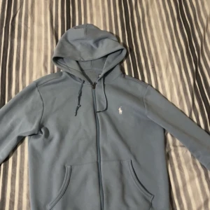 Blå hoodie från Ralph Lauren - Säljer en snygg blå hoodie från Ralph Lauren i storlek M. Den har en dragkedja framtill och en klassisk logga på bröstet. Perfekt för både höst och vår, med en skön passform och kängurufickor. Huvan har justerbara snören för extra komfort. Sänker pris i snabb affär