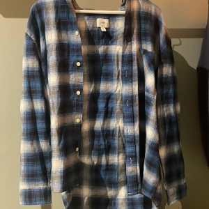 Blå och vit rutig flanellskjorta från H&M - Snygg blå och vit rutig skjorta från H&M i en avslappnad passform. Skjortan har långa ärmar och knappar framtill, perfekt för en casual look. Aldrig använd. Skjortan passar även M. Pris kan diskuteras.