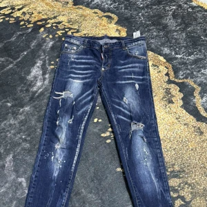 Blå jeans med slitningar - Säljer ett par blå jeans med coola slitningar och färgstänk. De har en klassisk femficksdesign och knappgylf. Perfekta för en avslappnad stil.