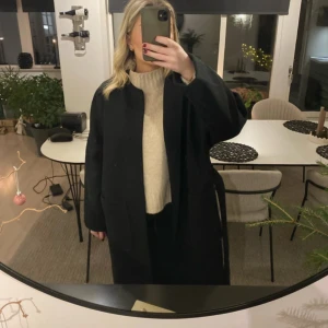 Svart kappa med bälte - Snygg svart kappa med bälte som ger en elegant look. Perfekt för kyligare dagar med sin långa design och klassiska stil. Passar bra över en stickad tröja för extra värme.