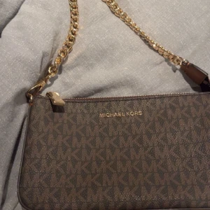 Brun axelväska från Michael Kors - Snygg brun axelväska från Michael Kors med kedjedetaljer och logotypmönster. Väskan har en elegant design med guldfärgade detaljer och en praktisk dragkedja. Perfekt för att ge din outfit en lyxig touch.