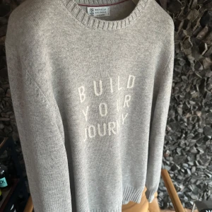 Grå stickad tröja från Brunello Cucinelli  - Säljer en stilren grå stickad tröja från Brunello Cucinelli med texten 'Build Your Journey' på framsidan. Tröjan har långa ärmar och en rund halsringning. Perfekt för kyliga dagar! Tillverkad i Italien med dem bästa tygen. 60% ull, 30% Kashmir, 10% silke. Skriv vid frågor.