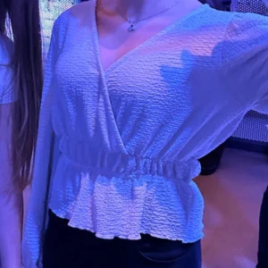 Vit omlottblus - Säljer en stilren vit omlottblus med v-ringning och långa ärmar. Blusen har en elastisk midja som ger en smickrande passform. Perfekt för både vardag och festliga tillfällen.