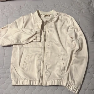 Vit silkes jacka - Snygg vit bomberjacka med dragkedja och ribbade muddar. Perfekt för en stilren look. Jackan har två fickor framtill och en klassisk bomberkrage.