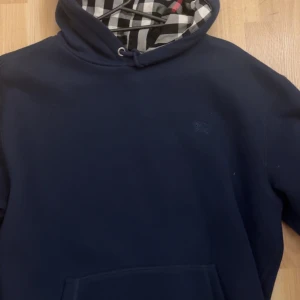Mörkblå hoodie från Burberry - Säljer en stilren mörkblå hoodie från Burberry med klassiskt rutigt mönster i luvan. Tillverkad i 100% bomull för en bekväm känsla. Perfekt för en avslappnad look.