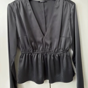 Grå blus från Gina Tricot - Snygg grå blus från Gina Tricot med v-ringning och långa ärmar. Blusen har en elastisk midja som ger en fin passform och ett lätt glansigt material som ger en elegant touch. Perfekt för både vardag och fest!