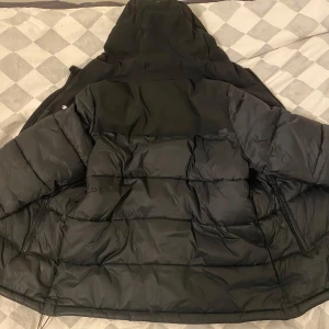 Svart pufferjacka från Champion - Säljer en svart pufferjacka från Champion med dragkedja och huva. Jackan har en logotyp på bröstet och är perfekt för kyliga dagar. Den är både stilren och funktionell.