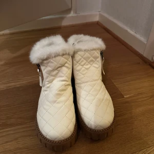 Vita quiltade boots med pälsdetalj - Vita quiltade boots med fluffig pälsdetalj runt kanten och dragkedja på sidan. De har en robust brun sula som ger bra grepp. Perfekta för kyliga dagar.