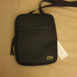 Svart axelväska från Lacoste - Snygg svart axelväska från Lacoste med det klassiska krokodilmärket. 500 kr off av mig. Väskan har en justerbar axelrem och en praktisk dragkedja framtill. Perfekt för att bära med sig det nödvändigaste.