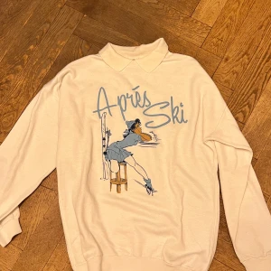 Vit tröja med 'Après Ski' tryck - Säljer en vit tröja med ett coolt 'Après Ski' tryck i blått. Tröjan har en krage och långa ärmar. Perfekt för en avslappnad stil med en touch av retro.