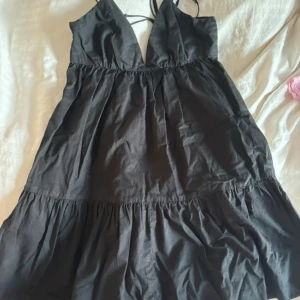 Svart klänning med v-ringning - Snygg svart klänning med v-ringad hals och knytband. Perfekt för en stilren look. Klänningen har en luftig passform och är ärmlös, vilket gör den bekväm och lätt att bära.