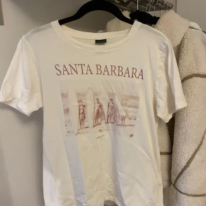 Vit t-shirt från Gina Tricot  - Säljer en vit t-shirt från Gina Tricot med trycket 'Santa Barbara' och en bild i rött. T-shirten är kortärmad och gjord i bomull, perfekt för en avslappnad stil.💓