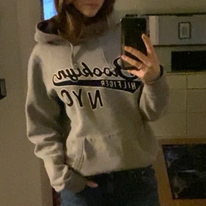 Grå hoodie från Hilfiger - Säljer en snygg grå hoodie från Hilfiger med texten 'Brooklyn NYC' på framsidan. Den har en klassisk känguruficka och justerbar huva. Perfekt för en avslappnad stil.