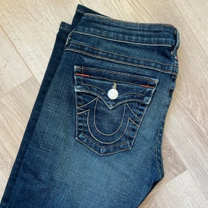 Mörkblå True Religion jeans  - Snygga mörkblå jeans från True Religion med unika broderade bakfickor och kontrastsömmar. De har en klassisk femficksdesign och knappgylf. Perfekta för en stilren look.