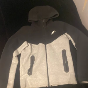 Nike tech fleece - Säljer en grå och svart Nike tech fleece. Tröjan har en klassisk Nike-logga på bröstet och är perfekt för en sportig look. Huvan ger extra komfort och stil. 