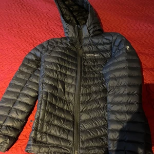 Svart dunjacka från Peak Performance - Säljer en svart dunjacka från Peak Performance med huva och dragkedja. Jackan är perfekt för kyliga dagar och har en stilren design med diskret logga på bröstet..