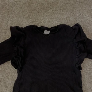 Svart ribbad tröja från Zara - Säljer en svart ribbad tröja från Zara med volangdetaljer längs ärmarna. Perfekt för en stilren look med det lilla extra.🤍