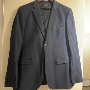 Mörkblå kostym - Elegant mörkblå kostym med kavaj och matchande byxor. Kavajen har två knappar framtill och fyra dekorativa knappar vid ärmsluten. Perfekt för formella tillfällen.