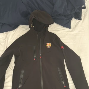 Svart jacka från Barca  - Snygg svart jacka med FCB-logga på bröstet och texten 'BARÇA' på ärmen. Jackan har en dragkedja framtill och en huva för extra skydd. Perfekt för fotbollsfans som vill visa sitt stöd. Köpt i camp nou för 2000