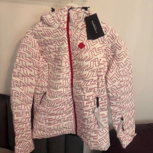 Vit och röd skidjacka från J.Lindeberg - Snygg vit skidjacka med rött mönster från J.Lindeberg. Jackan har en dragkedja framtill och en justerbar huva. Perfekt för kyligare dagar med sin stilrena design och praktiska fickor. Säljer även matchande skidbyxor. Helt oanvänd med prislapp