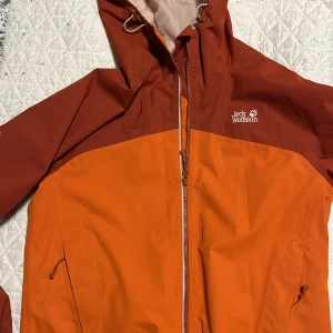 Orange och röd jacka från Jack Wolfskin - Säljer en funktionell orange och röd jacka från Jack Wolfskin. Jackan har en praktisk huva och dragkedja framtill. Perfekt för utomhusaktiviteter med sin tåliga design. Finns en färgfläck enligt bild 4.