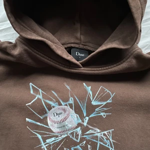 Dime hoodie - Säljer en brun hoodie från Dime med ett coolt tryck av en baseboll som krossar glas. Trycket är lite tvättat men ser vintage ut. Hoodien har en  känguruficka och en bekväm passform. 