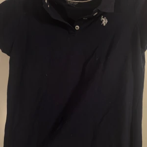 Mörkblå pikétröja från U.S. Polo Assn. - Säljer en stilren mörkblå pikétröja från U.S. Polo Assn. med korta ärmar och klassisk krage. Tröjan har en broderad logga på bröstet och knappar framtill. Perfekt för en avslappnad men ändå snygg look.