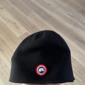 Canada goose mössa  - Snygg svart mössa med en rund logotyp framtill. Perfekt för kyliga dagar och enkel att matcha med olika outfits.