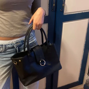Svart handväska  - Snygg svart handväska med stilren design. Väskan har en  ringdetalj framtill och två bekväma handtag. Perfekt för att bära dina dagliga nödvändigheter❤️❤️💕