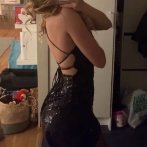 Svart glitterklänning - Snygg svart klänning med paljetter som glittrar i ljuset. Den är tight och kort, perfekt för fest eller nyår. Axelbandslös design med tunna band som korsar på ryggen för en extra touch. Klänningen är i mycket bra skick och redo att bäras för en glamorös kväll!