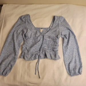 Blommig blå blus med volangdetaljer - Söt blommig blå blus med långa ärmar och volangdetaljer. Blusen har en knytning framtill och en elastisk rygg för en bekväm passform. Perfekt för vår och sommar!