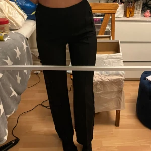 Svarta byxor med hög midja - Snygga svarta kostymbyxor med hög midja och rak passform. Perfekta för en stilren look. Byxorna har en diskret glansig detalj längs sidorna som ger en extra touch. Resår i midjan så sköna upptill. 