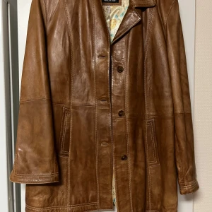 Brun skinnjacka från Goldstein - En snygg lite längre brun skinnjacka från Goldstein med fina detaljer. Jackan har mönstrad insida i ljusa färger. Den ger lite vintage vibe och är perfekt för höst och vår. 